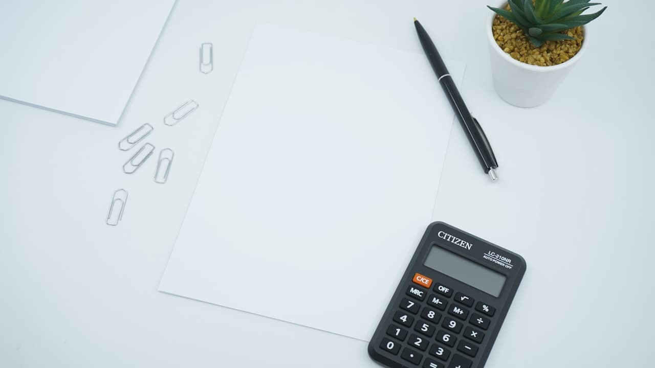 A bright white desk with a blank sheet of paper, paper clips, a pen, a calculator, and a small potted plant.